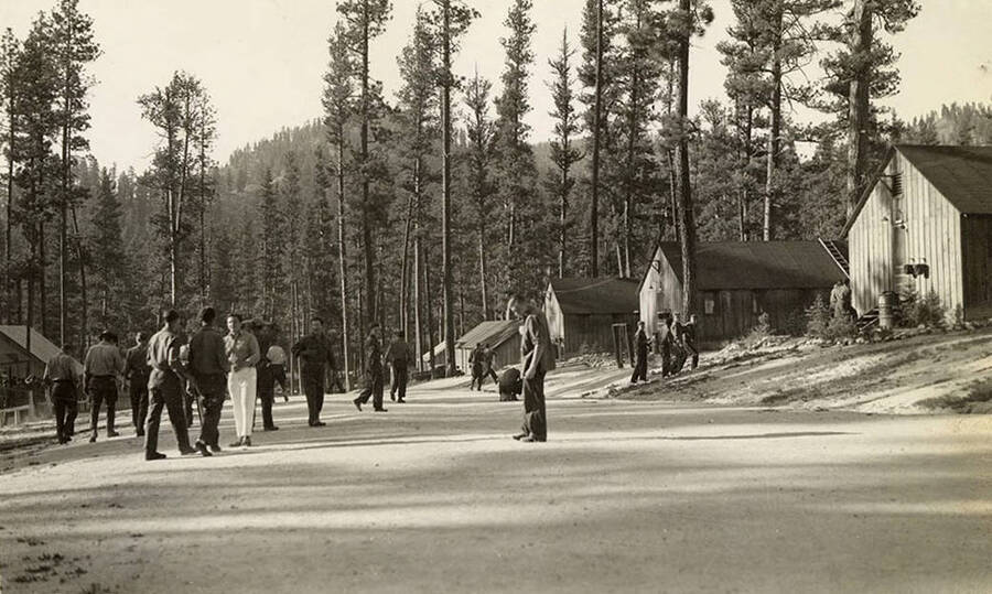 The CCC men rest in camp after dinner. Here they are seen standing around in the road between buildings and barracks. Writing under the photo reads: 'After Chow'.