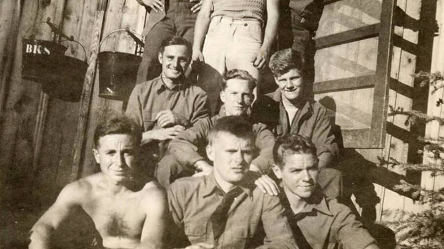 Group of CCC men posed on the front steps of their barrack. Six men are sitting and three are standing, but their upper bodies are cut out of the photo. Letters on the bucket hanging on the wall reads: 'BKS'. Writing under the photo reads: 'Some of the boys'.
