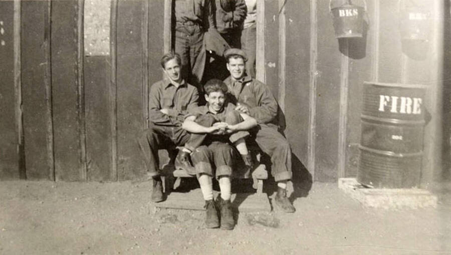 Group of CCC men posed on the front steps of their barrack. Three men are sitting, three more are standing but their upper bodies are cut out of the photo. Letters on the buckets hanging on the wall of the barrack read: 'BKS' Writing on the barrel reads: 'Fire'. Writing under the photo reads: 'Before the work call'.