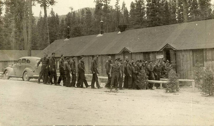 A company of CCC men line up for dinner outside a building with a sign that reads: 'Mess Hall'. Writing under the photo reads: 'Come and get it'.