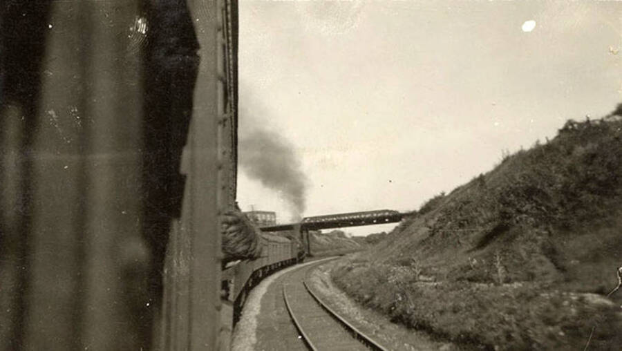 A CCC man sticks his head out of the train for a look at the railroad tracks and bridge crossing above the train ahead.