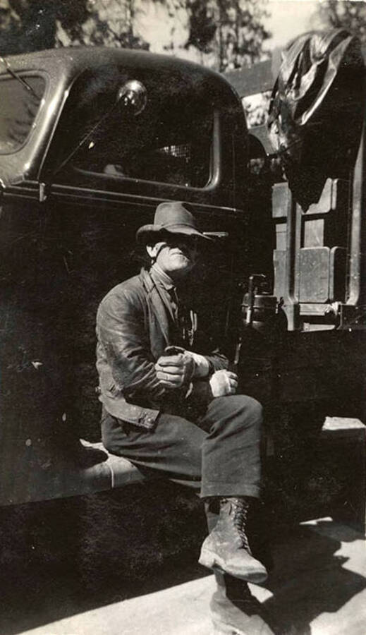 CCC man (possibly an LEM or foreman) sitting on the side of a truck, his hat shading his eyes. Writing under the photo reads: 'John'.