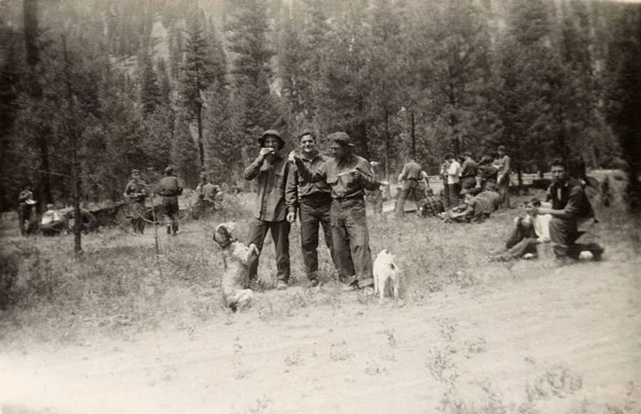 A CCC work crew having lunch at the job site. In the center of the photo are three CCC men standing in a line, while two dogs beg for food at their feet. The other CCC men are sitting or standing in the background. Writing under the photo reads: 'Chow time on Airport'.