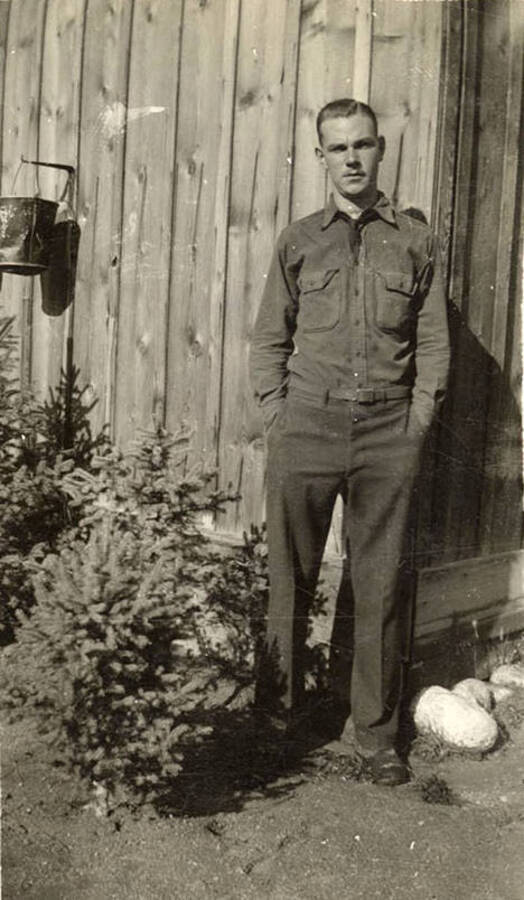 Photo of a CCC man posed at the corner of a building, small pine trees grow to his right. Writing under the photo reads: 'Me'.
