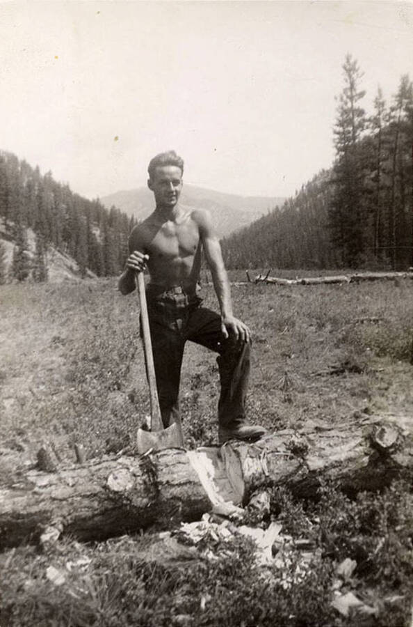 A CCC man posing with an axe chopping a log in the middle of an airfield under construction. Writing under the photo reads: 'Geurge McMann'.