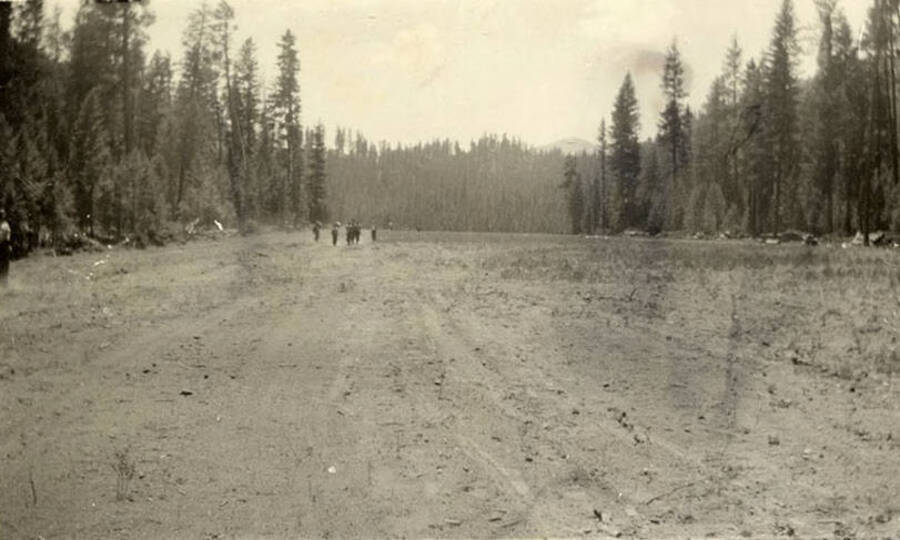 Several CCC men walking across an airfield in the distance. Both sides of the field are lined with trees and a wooded hill can be seen in the far background. Writing under the photo reads: 'Back to work'.