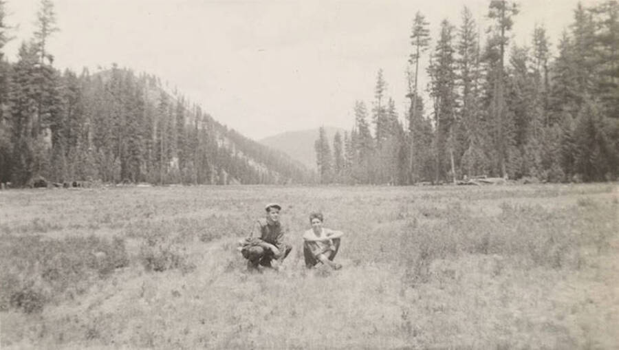 Two CCC men sitting in an airfield, forested hills rise in the background. Writing under the photo reads: ''Two-stips' and Lotte'.