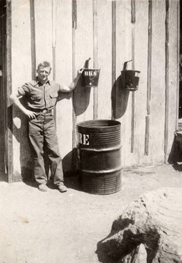 Photo of a CCC man standing outside a barrack at CCC camp F-168. He is standing next to a barrel and holding a bucket on the wall that reads: 'BKS'. Writing under the photo reads: 'Mazak'.