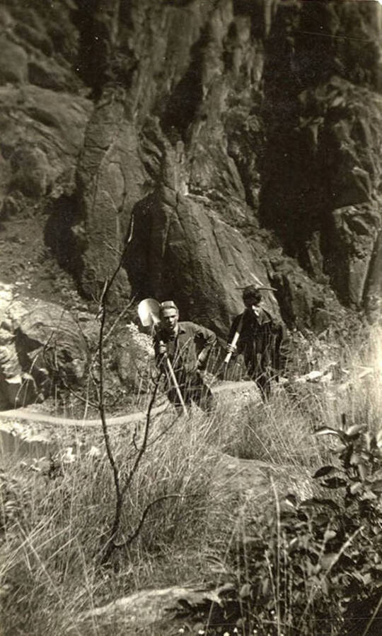 Two CCC men, one holding a pick axe, the other a shovel, pose on the edge of a river canyon, the ground falling away behind them, the river barely visible, and a tall cliff on the other side of the bank.