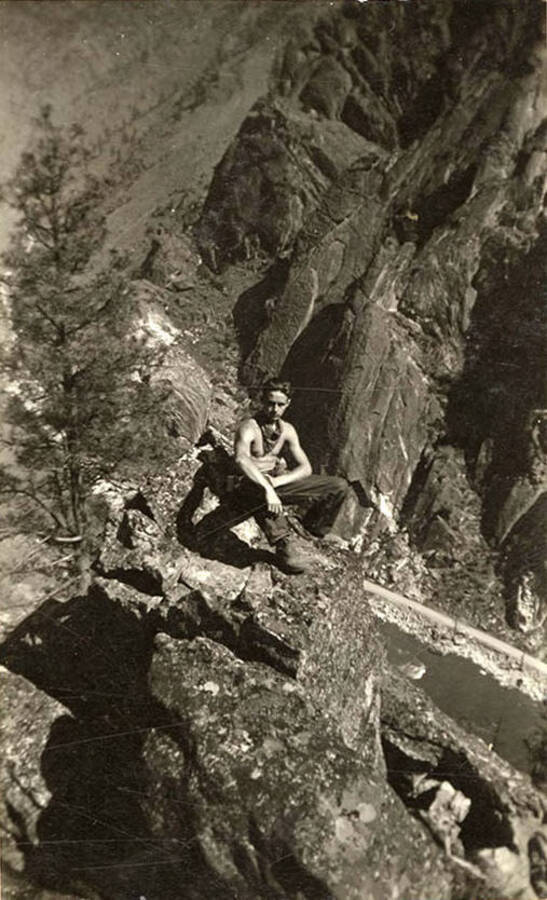 Photo of a CCC man posing on the edge of a river canyon. The river can be seen far below and a tall cliff face rises on the other side of the bank.
