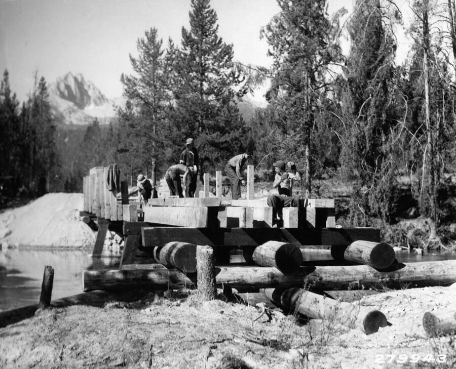 Several CCC men working on a bridge building project near Redfish Lake in the Sawtooth National Forest. One of the Sawtooth Mountains can be seen in the background. Back of the photo reads: 'U.S. Forest Service Photo. Bridge building near Redfish Lake, Sawtooth National Forest, Idaho. Taken by K.D. Swan 1933.'
