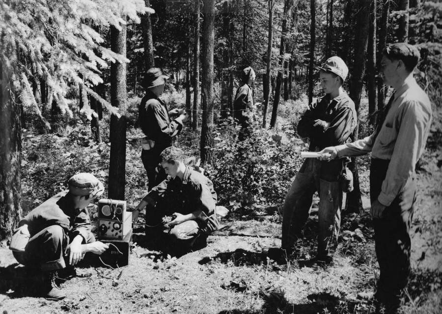 Several CCC men participate in a National Defense training in the woods. The training is given by the McCall Radio School and the U.S. Office of Education, near Camp Lake Fork, Company 2939. Writing above photo reads: 'Camp Lake Fork Company #2939, McCall, Idaho July, 1941. National Defense Training - McCall Radio School - The radio instructor of the McCall radio school, McCall Idaho, has a group of CCC enrollees out in the nearby forest demonstrating how to unpack, set up, and operate a Forest Service SPF radio set. This is but one of the many features stressed in an intensive six month radio training course sponsored by the U.S. Forest Service. Technical service supplied by the State of Idaho and the U.S. Office of Education, National Defense Training. Photo by W.J. Mead.'
