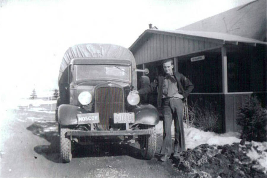 A CCC man leans against the hood of a covered truck on a road in front of a building in a CCC Camp. The front of the truck has two plates on it. One of them reads: 'Moscow', the other is a license plate: '16846'. There is a pile of snow on the side of the road. Writing under the photo reads: 'CCC Camp Moscow, Idaho 1938-1939'.