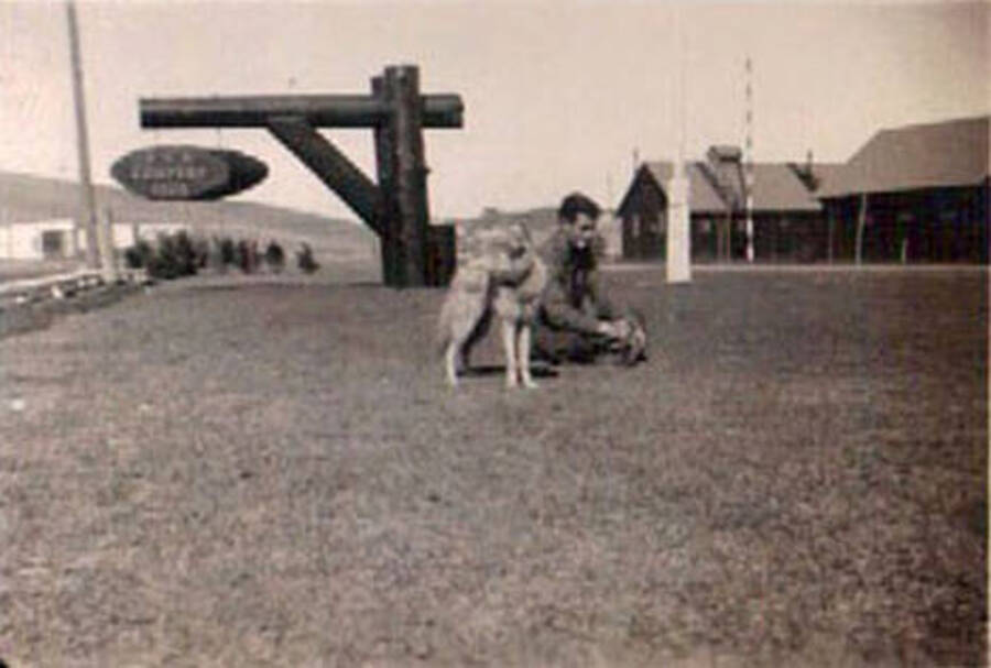 A CCC man kneels next to a canid on the lawn of a CCC Camp. The animal is either a dog, wolf, or coyote. There is a sign in the background, but it is illegible.