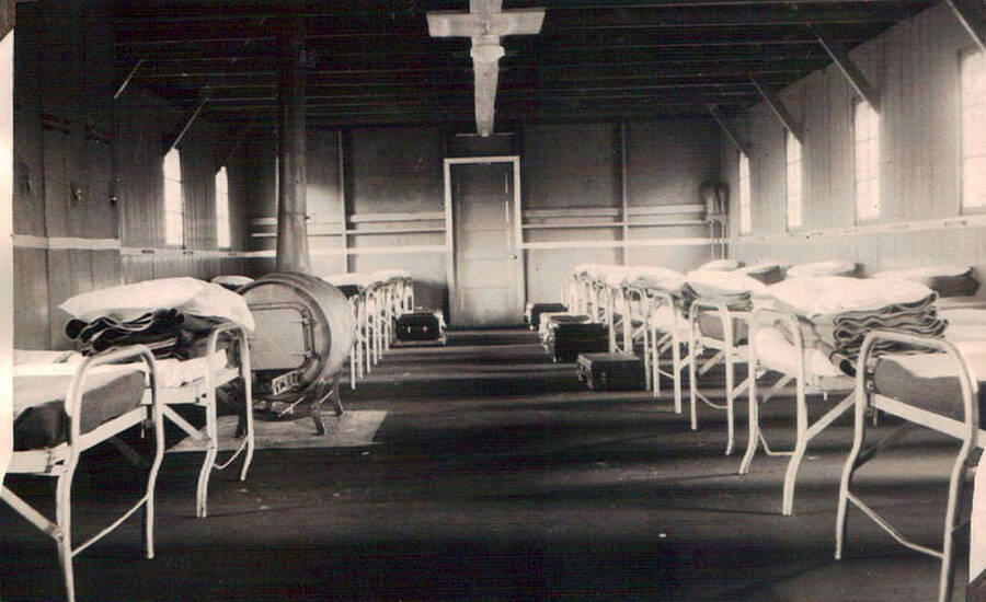 Photo of the inside of barracks with beds and barrel stove at a CCC camp. A pile of folded blankets sits at the end of each bed. Some beds at the end near the door have trunks at the foot of the bed. Writing above the photo reads: 'K.P. Barracks'