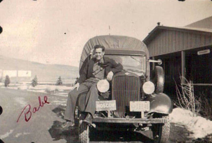 A CCC man sits on top of a covered truck on a road in front of a building at a CCC Camp in Moscow, ID. Two plates are on the front of the truck, one reads: 'Moscow' the other is a license plate: '16845'. Writing on the album page reads: 'Lewiston, Idaho 1938 CCC Camp'. Writing under the photo reads: 'camp'. Writing on the photo reads: 'Babe'.