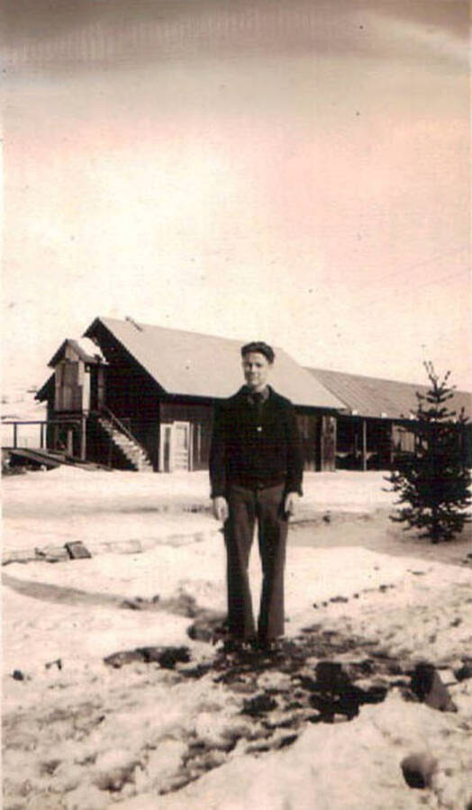 Photo of a CCC enrollee standing in a snowy CCC camp with a building in the background.