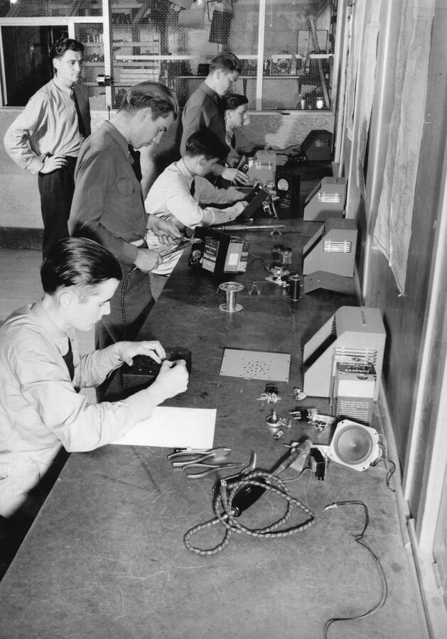 Several CCC men participate in a National Defense training on how to build radios. The training is given by the U.S. Office of Education and the McCall Radio School near CCC Camp Lake Fork, company 2939. Taken in July. The writing above and on the back of the photo reads: 'Camp Lake Fork Company #2939, McCall, Idaho July 1941. National Defense Training -McCall Radio School- Unit Radio Production - Here are a group of CCC enrollees attending classes at the McCall, Idaho national defense radio school. Students in the class are being shown how to produce and build their own radio oscillators for code practice. Each man learns to assemble a specific part of the set. The enrollee in the front is laying out the front panel which he will drill for the parts which the second is wiring. The third enrollee is making a check on the different (over) (Cont'd.) instruments before the fourth one finally places the panel in its metal case. Final check-up and test is made by the enrollee at the end. This training, on a smaller scale, is given to enrollees to adapt them, if need be, to small scale radio set production in factories. Photo by W.J. Mead.'