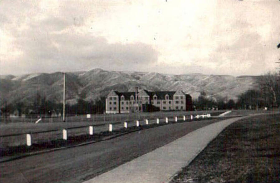 A scenic photo of a large building surrounded by fenced fields and roads with a sidewalk. There are also a range of hills in the background. Writing on the album page reads: 'Lewiston, Idaho 1938 CCC Camp'. Writing under the photo reads: 'Lewiston Idaho'.