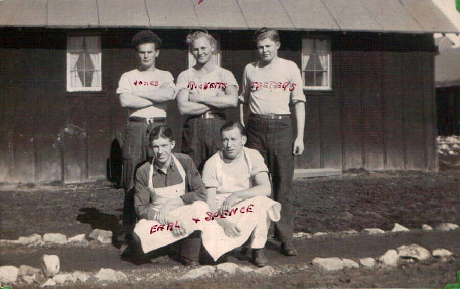 Group photo of a kitchen crew in front of a building in a CCC camp. Writing above the photo reads: 'Mercer's Shift'. Names of each cook are written across the photo in red ink: 'Jones, Ricketts, Teeters, Earl and Spence'.