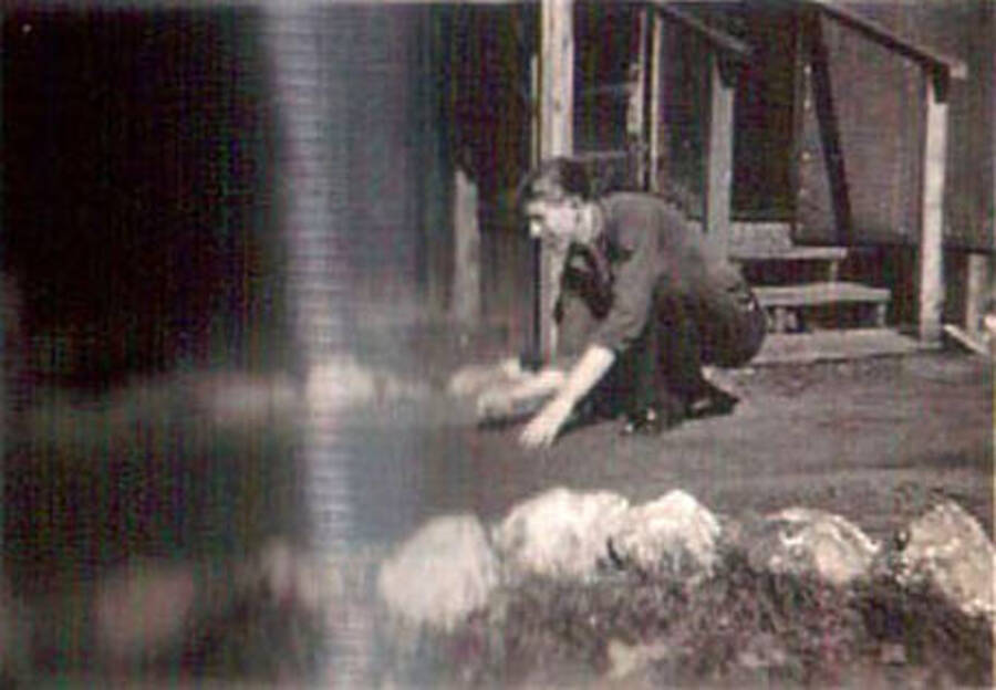 A CCC enrollee kneels on a path in front of a building in a CCC camp. The left half of the photo is blurred.