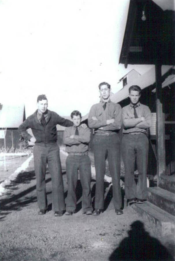 A group of four CCC men pose for a picture in the Moscow, ID CCC Camp. Writing under the photo reads: 'CCC Camp Moscow, Idaho 1938-1939'. An arrow pointing to the shadow in the bottom right hand corner is labeled: 'Johnny'.