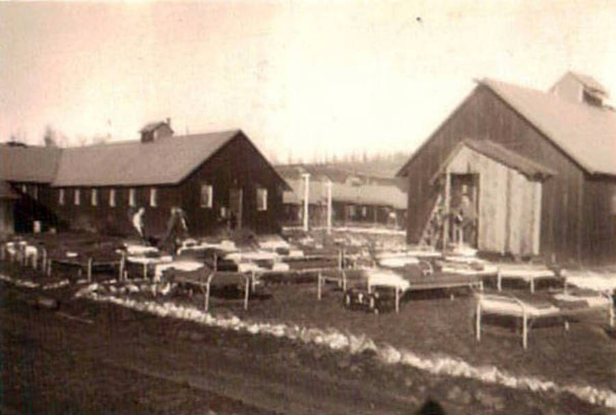 A view of a CCC camp with cots set out on the lawn in front of the barracks.