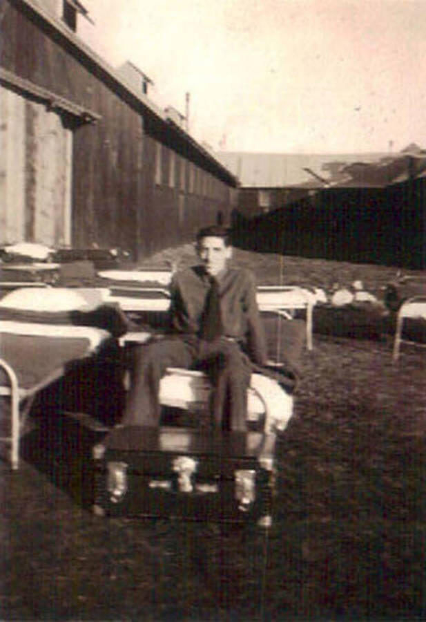 A CCC man poses on a cot on the lawn of a CCC camp, there are other cots on the lawn and a trunk sitting at the end of his. Writing to the left of the photo reads: 'Spahgetti'.