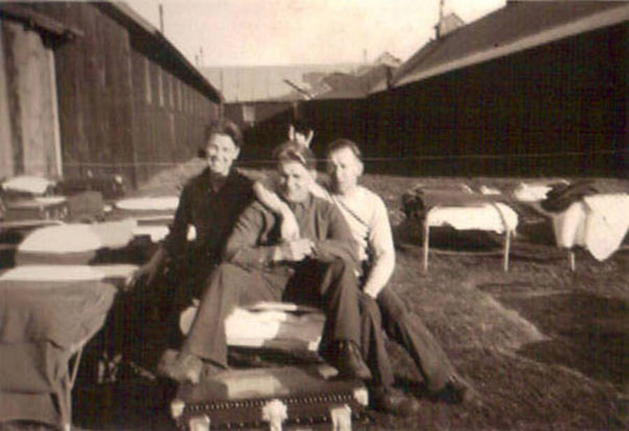 Three CCC men pose on a cot on the lawn of a CCC camp. There are other cots on the lawn and a trunk at the foot of the cot the men are sitting on.