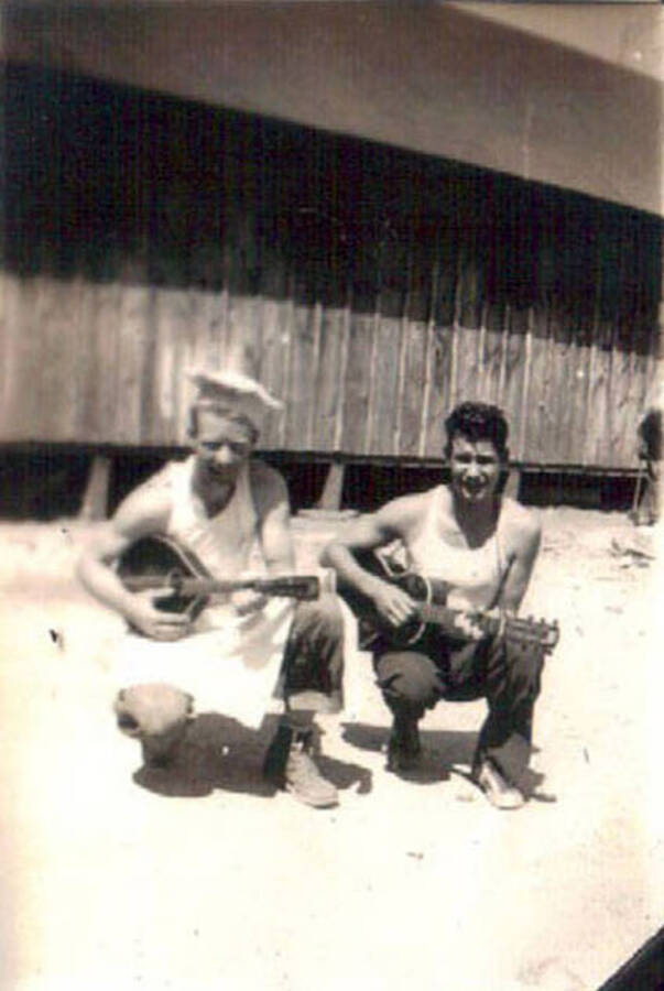 Two CCC men kneeling next to a CCC barrack holding two stringed musical instruments. One appears to be a mandolin, the other a small guitar. Label on the album page reads: 'John's friends Idaho'.