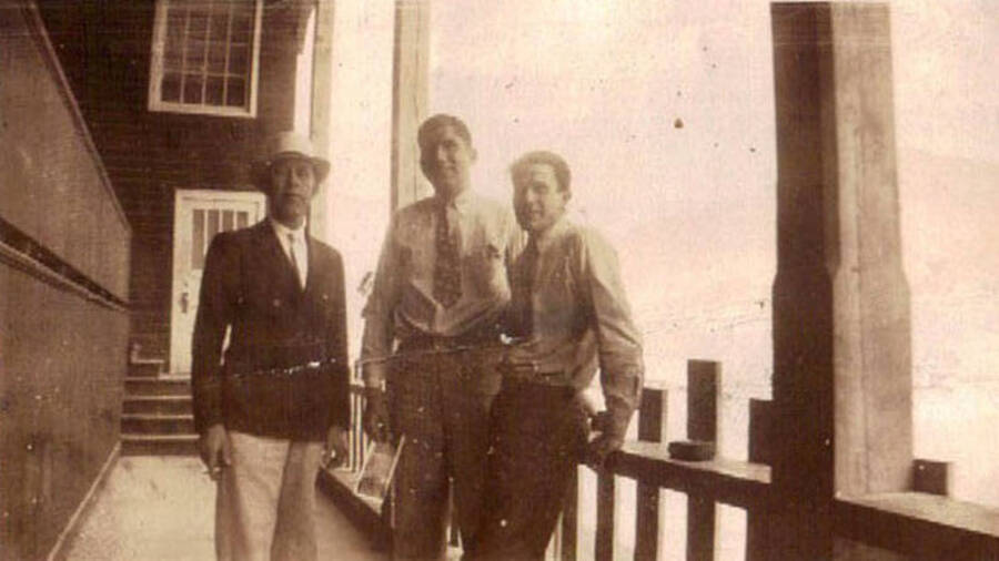 Three men lean against a fence in front of a building. The man on the left is wearing a jacket and dress hat. Label on the album page reads: 'John's friends Idaho'.