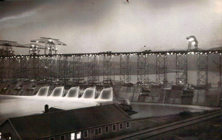 Night shot of Grand Coulee dam in Washington. The dam is topped by scaffolding and spotlights. Writing above the photo reads: 'Grand Coulee Dam Wash 1938'