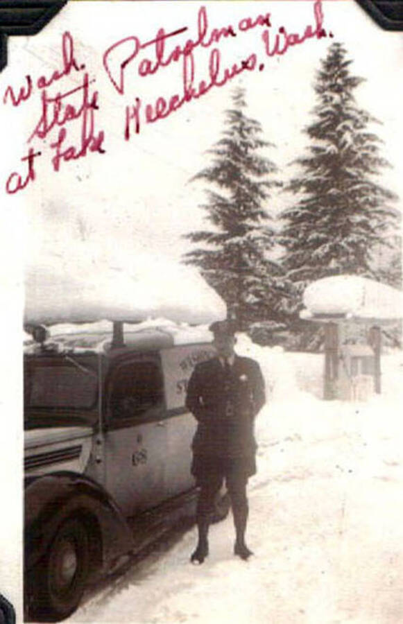 A uniformed officer in the snow standing next to his truck. Writing on the photo reads: 'Wash State Patrolman at Lake Keechelus, Wash'.