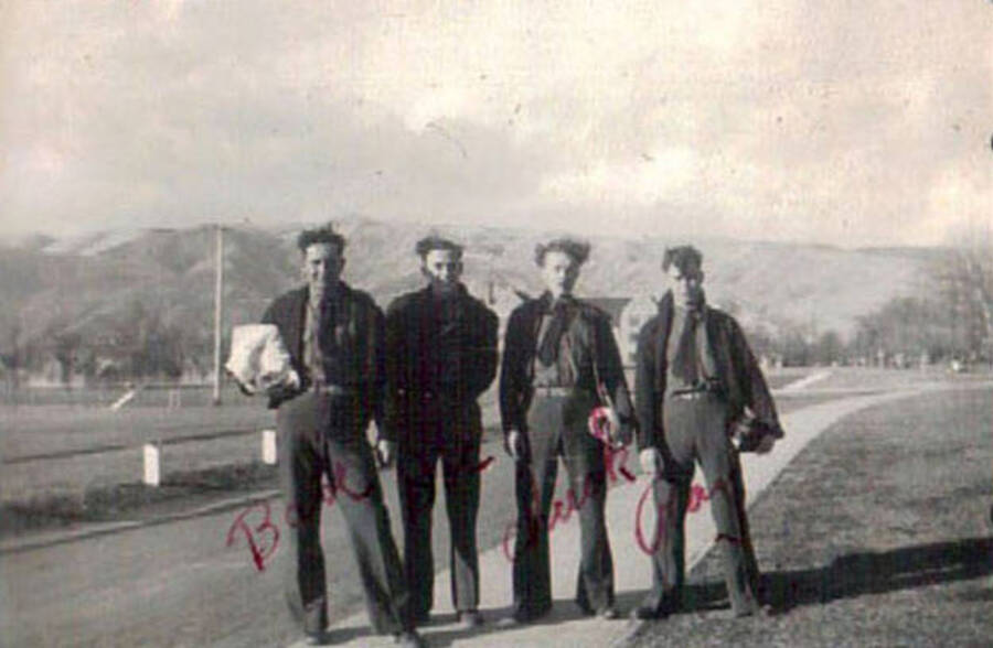 Four CCC men posing in front of a building, fenced lawns, roads, and range of hills. Writing beneath the photo reads: 'Lewiston Gym'. Writing on the photo includes each man's name.