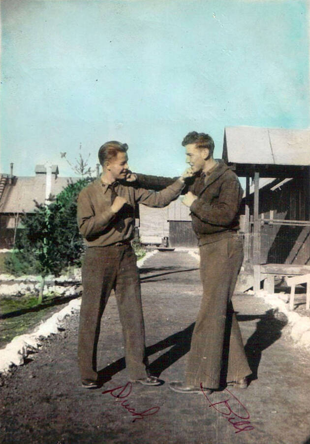 A photo of two CCC men posing for a photo in the Moscow, Idaho CCC camp. The photo has been colored. John and Bill; color photograph. Writing on the photo labels each man as 'Duck' and 'Bill' from left to right. Writing under the photo reads: 'Johnny 1938-39 CCC - Camp'.