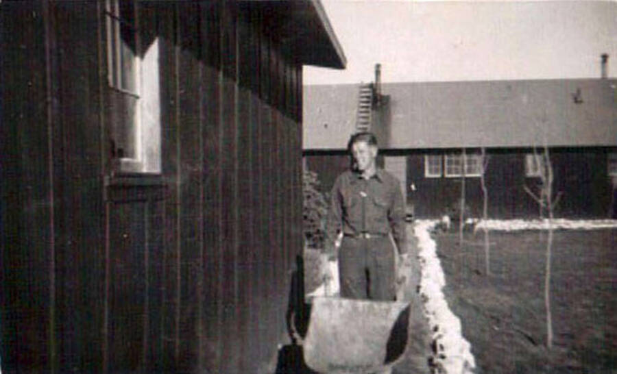 CCC man rolling a wheelbarrow down a path next to a barrack at CCC camp in Moscow, Idaho. Writing under the photo reads: 'Slim Nelson'.