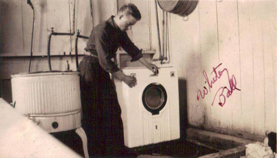 A CCC man stands by next to a washing machine in a CCC camp in Moscow, Idaho. Writing on the photo reads: 'Whitey Ball'.