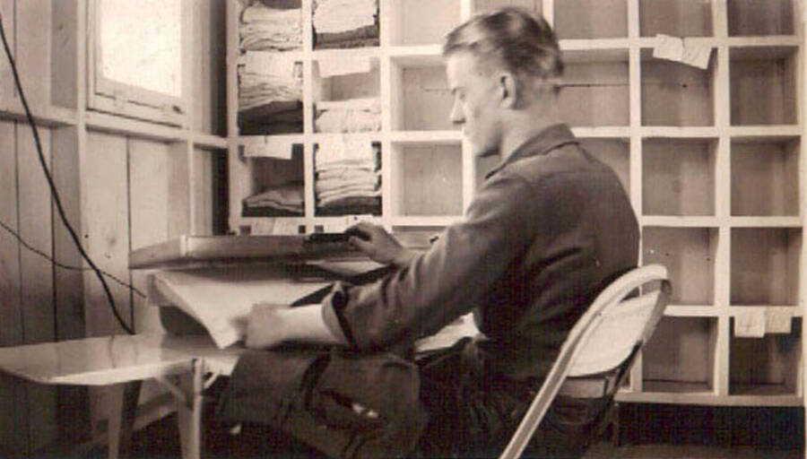 A CCC man uses a clothing press to iron a pair of pants. A wall of cubby holes stands behind him, some of the cubbies are full of folded clothing, most are empty.