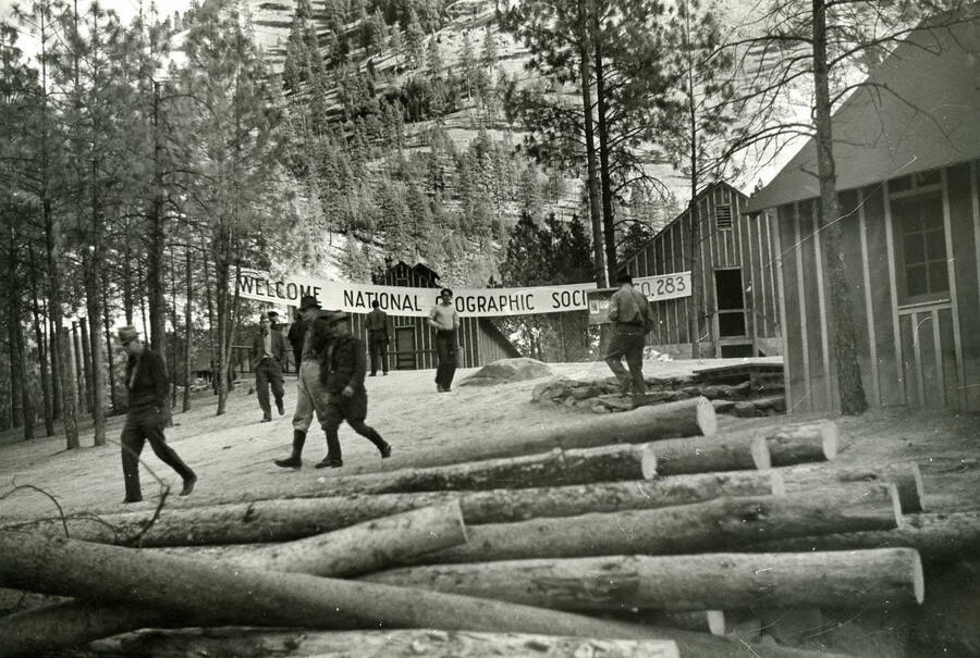 A sign hangs between trees and in font of CCC buildings, welcoming the National Geographic expedition visitors to CCC Camp F-109. Several men are walking and standing around the camp and a pile of logs lays in the foreground of the picture. The banner reads: 'Welcome National [Ge]ographic Soc[iety] [C]o. 283'.