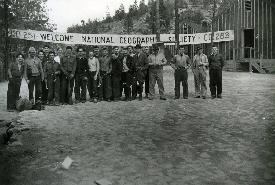A group of CCC men and staff lined up under a welcome sign for the National Geographic Society. The banner reads: 'Co. 251 Welcome National Geograph[ic] S[oc]iety Co. 283'.