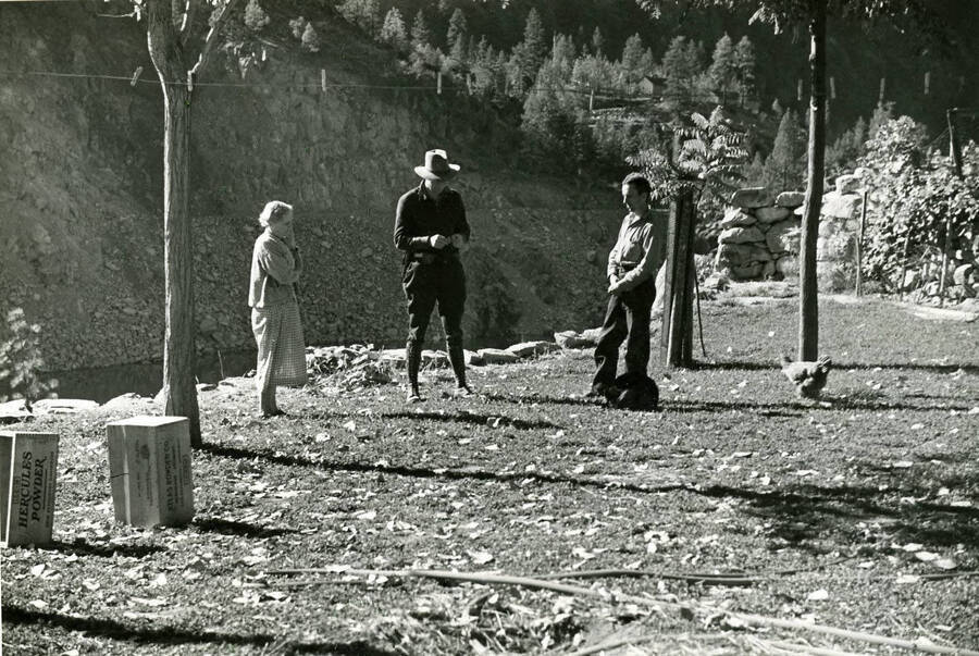 A photo of three individuals standing in Mrs. Standish's lawn across Salmon River from Salmon River Road. There is also a chicken, two upright wooden boxes, and a clothes line in the yard. Writing on the box reads: 'Hercules powder'.