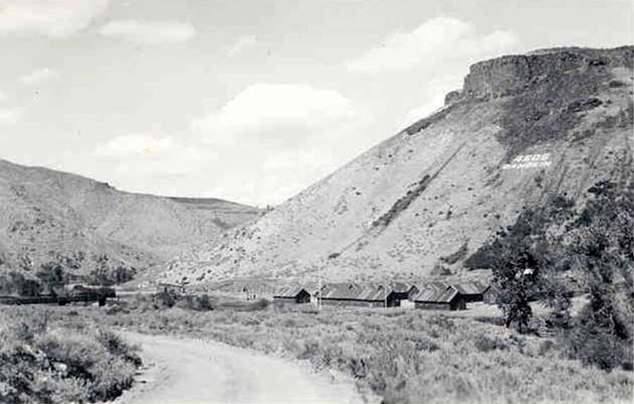 Overview of Danskin CCC Camp and the surrounding area. There is a road in the foreground and hills in the background. There is also writing on the hill behind the camp.