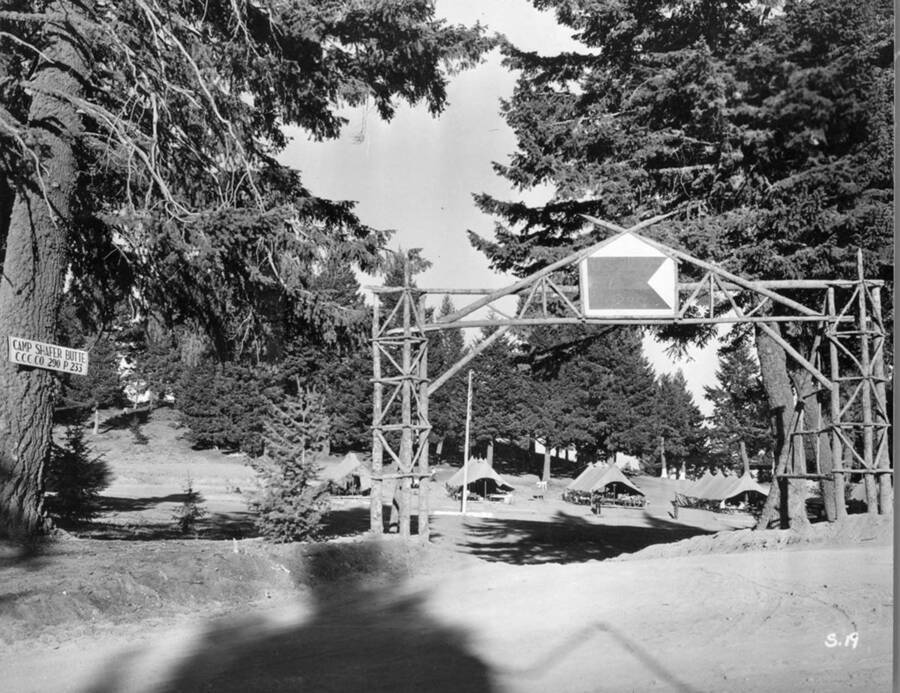 Road and entrance to Shafer Butte CCC Camp. Tent barracks can be seen in the background. A sign posted to a tree on the left side of the photo reads: 'Camp Shafer Butte CCC Co 290 P.233'. The sign above the entrance reads: 'U.S. Co. 290'.