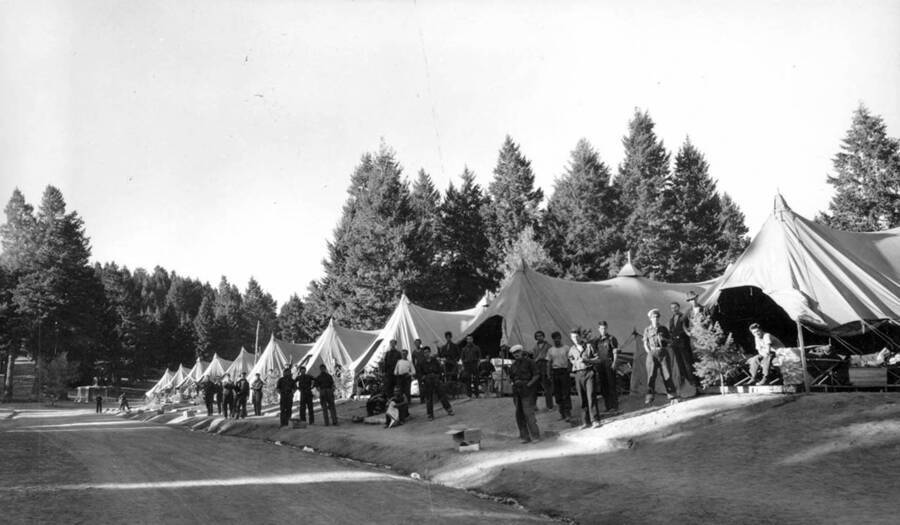 Groups of CCC men stand outside their tent barracks at Shafter Butte CCC Camp. The barracks are lined up along one side of a road and trees can be seen rising behind the tents in the background.