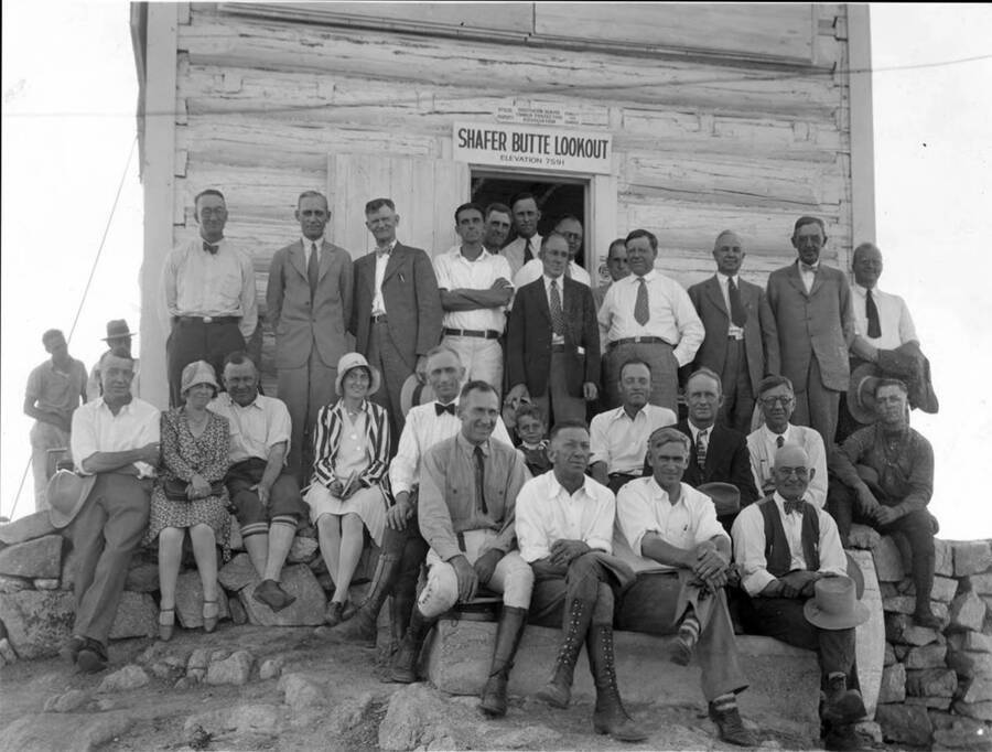 A large groupe of older men, two women and one child poses for a photo in front of Shafer Butte Lookout. The sign above the door to the lookout reads: 'Shafer Butte Lookout Elavation 7591'. The sign above that is hard to read but part of the sign reads: 'Southern Idaho Timber Protective Association'.