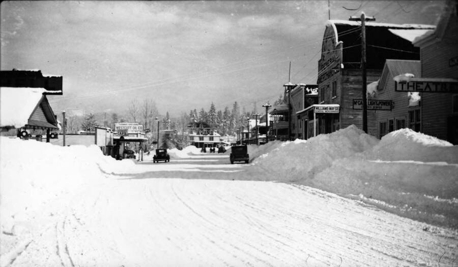 View of a snow-covered street in downtown McCall. Writing on the photo reads: '1850.0005. CCC McCall - facing east Author: David Lyon 1935/36 Print (from original negative)'. Writing on various signs in the photo reads: 'Goodman's Super Service Westcott Oil Co.' 'Ford' 'Goodman's Cafe' 'Murphy Hotel' 'Coffee Sho[p]' 'N. [F.] Williams General Merchandise' 'Williams May Co. General Merchandise' 'McCa[ll] Light & Power E[lec]tric Service' 'Theatre'