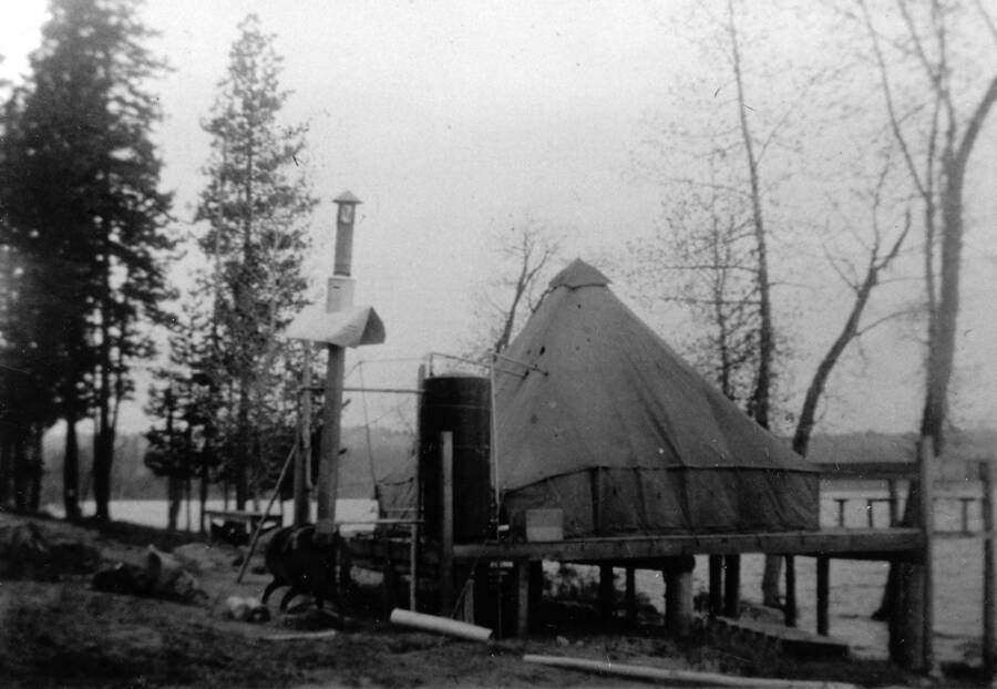 Photo of a tent on a platform above the edge of Payette Lake. Writing beneath the photo reads: '1850.0008. CCC Camp S-223 McCall, ID 1935 Shower Tent Erected on Platform of Old Camp. Author: David Lyon Print (x2) (Negative in '1850 D. Lyon Negatives') 3A & 21 A Color'.