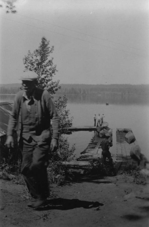 Photo of a man walking up the hill away from the lake with other men standing on piers in Payette Lake in the background. Writing beneath the photo reads: '1850.0009. CCC CCC Camp S-223  McCall, ID George Newcomb, carpenter, helped build camp Author: David Lyon Print (x2) (Negative in '1850 D. Lyon Negatives') OA & 17A Color'.
