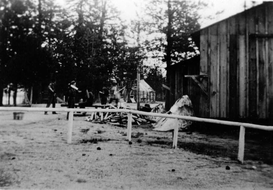 CCC crew chopping firewood in front of the mess hall at CCC Camp S-223. Writing beneath the photo reads: '1850.0010. CCC Camp S-223 McCall, ID 1935 Firewood crew working in front of messhall. Author: David Lyon Print (Negative in '1850 D. Lyon Negatives') 4A Color'.