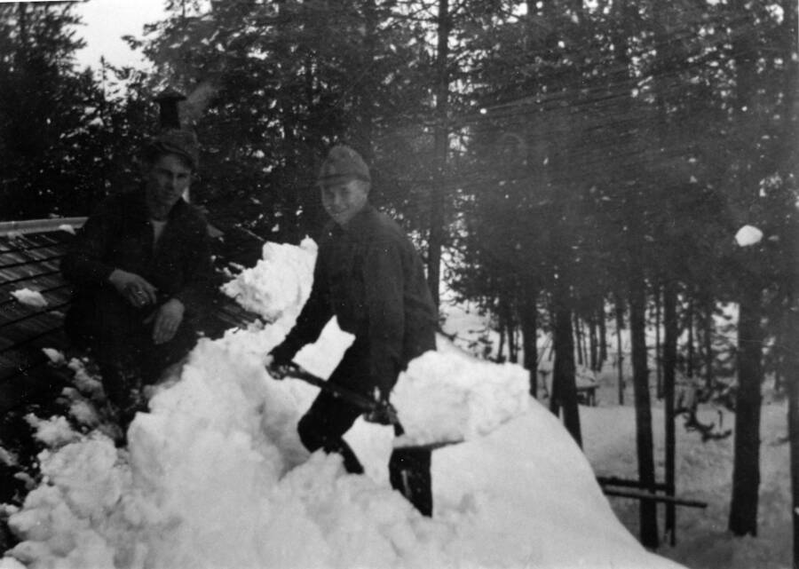 Two CCC men pose on a roof in the CCC camp. One of them holds a shovel full of snow. Writing beneath the photo reads: '1850.0015. Shoveling snow from roof of barracks, Cam S-223, McCall'